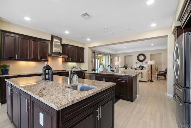 kitchen featuring appliances with stainless steel finishes, sink, light stone counters, a center island with sink, and wall chimney exhaust hood