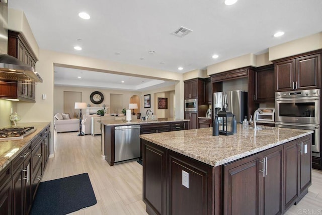 kitchen with appliances with stainless steel finishes, a kitchen island with sink, light stone countertops, dark brown cabinets, and wall chimney exhaust hood