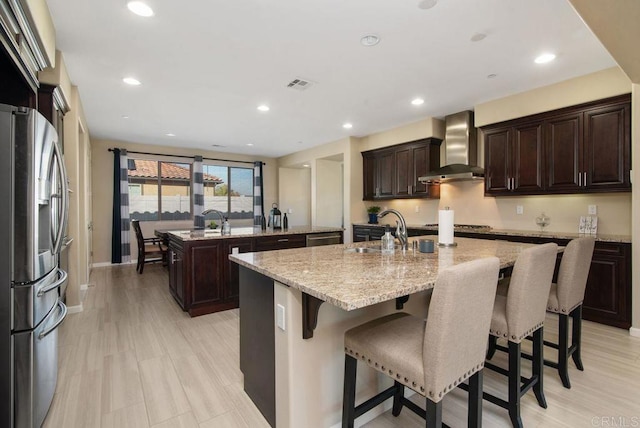 kitchen featuring wall chimney exhaust hood, sink, light stone counters, an island with sink, and stainless steel appliances