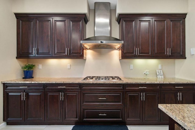 kitchen with stainless steel gas stovetop, light stone countertops, dark brown cabinets, and wall chimney range hood