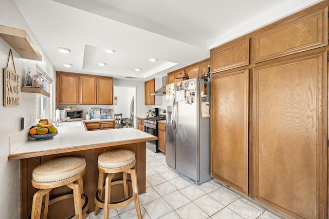 kitchen with appliances with stainless steel finishes, a kitchen bar, tile counters, kitchen peninsula, and a raised ceiling