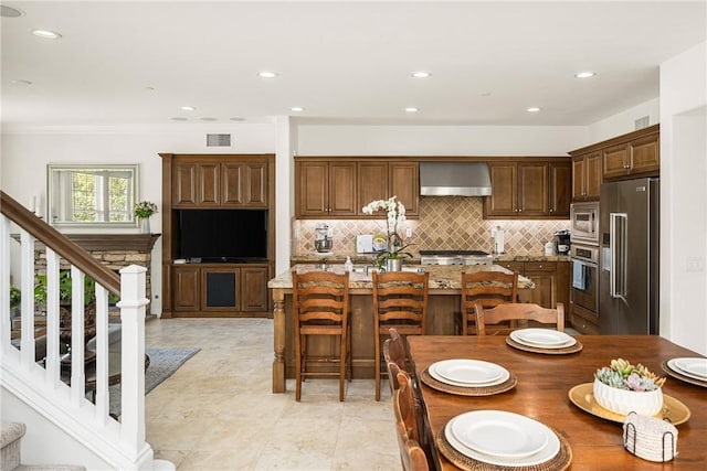 kitchen featuring light stone counters, stainless steel appliances, visible vents, an island with sink, and wall chimney exhaust hood