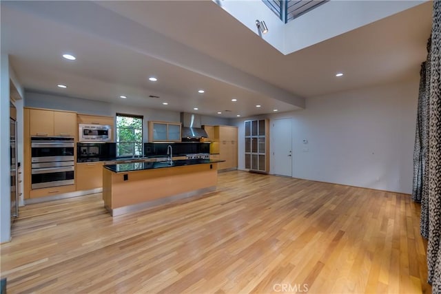 kitchen with a skylight, an island with sink, wall chimney exhaust hood, stainless steel appliances, and light hardwood / wood-style flooring