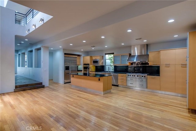 kitchen with wall chimney range hood, a kitchen island with sink, built in appliances, light wood-type flooring, and light brown cabinets