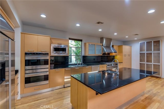 kitchen featuring appliances with stainless steel finishes, sink, a kitchen island with sink, wall chimney range hood, and light wood-type flooring