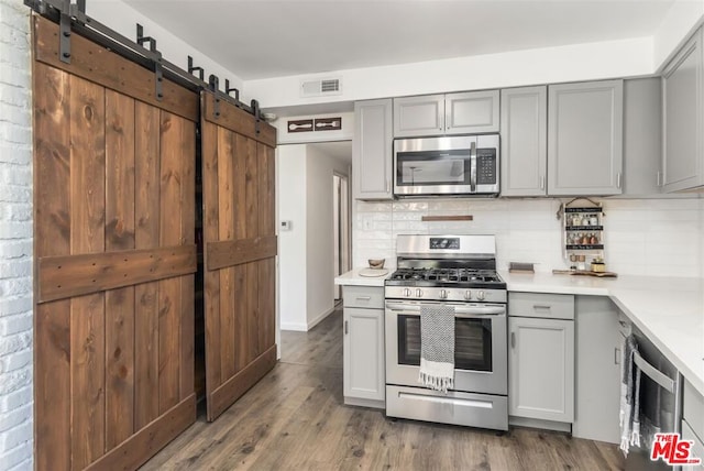 kitchen with gray cabinets, dark hardwood / wood-style flooring, decorative backsplash, stainless steel appliances, and a barn door