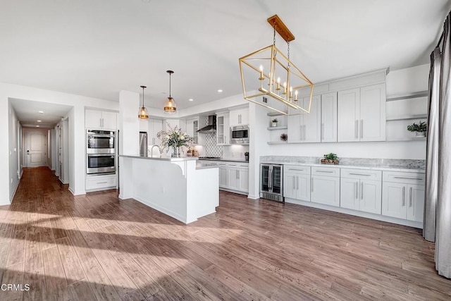 kitchen with pendant lighting, wall chimney range hood, appliances with stainless steel finishes, an island with sink, and white cabinets