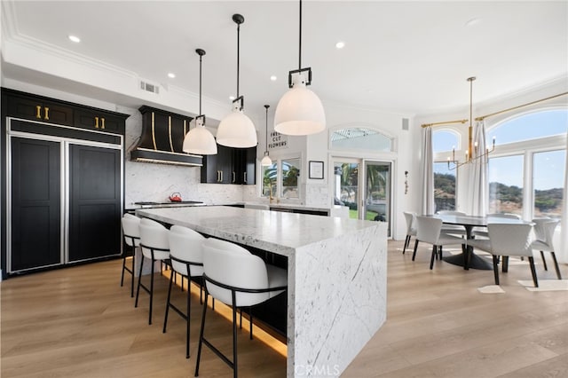 kitchen with a kitchen island, backsplash, hanging light fixtures, light stone counters, and custom range hood