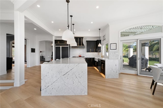 kitchen with a kitchen island, hanging light fixtures, paneled built in fridge, light stone countertops, and wall chimney range hood