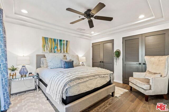 bedroom with multiple closets, wood-type flooring, a raised ceiling, and ceiling fan