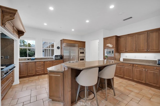 kitchen featuring sink, a kitchen bar, a kitchen island with sink, built in appliances, and light stone counters