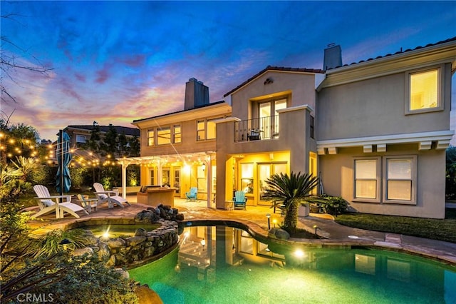 back of house at dusk featuring a fire pit, a patio area, a chimney, stucco siding, and a balcony