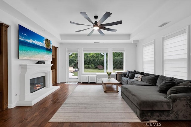 living area with visible vents, a ceiling fan, a raised ceiling, dark wood-style floors, and a glass covered fireplace