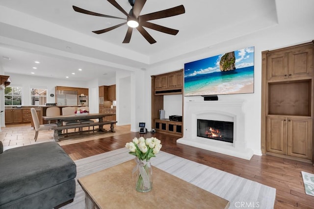 living room featuring a raised ceiling, a glass covered fireplace, light wood-style flooring, ceiling fan, and recessed lighting