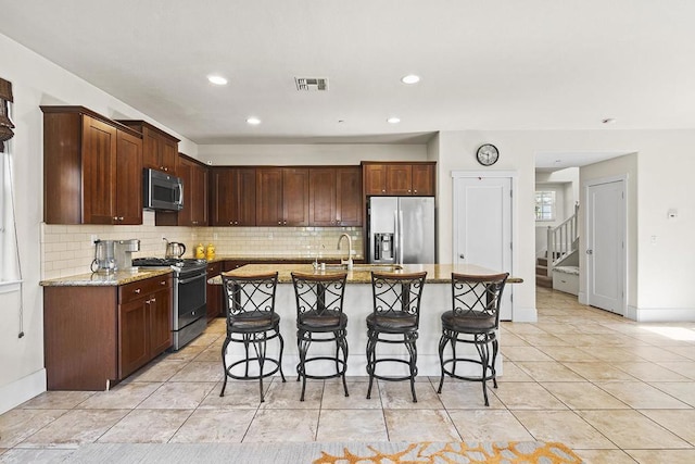 kitchen featuring a breakfast bar, light stone counters, tasteful backsplash, a center island with sink, and appliances with stainless steel finishes