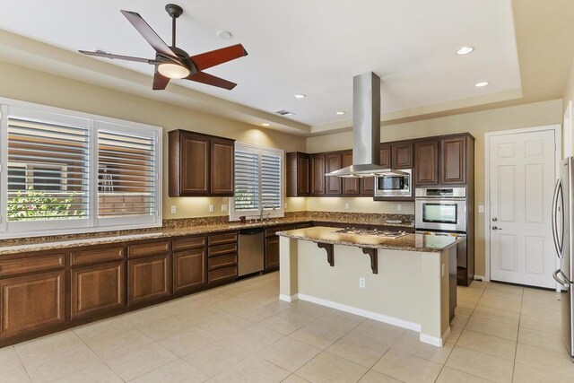 kitchen featuring a breakfast bar area, island range hood, appliances with stainless steel finishes, a kitchen island, and dark stone counters