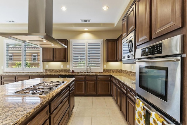 kitchen with sink, light tile patterned floors, appliances with stainless steel finishes, light stone counters, and island range hood