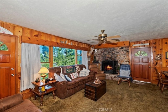 living room with a stone fireplace, wooden walls, ceiling fan, dark carpet, and a textured ceiling