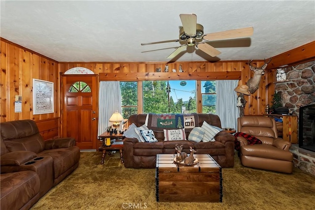 carpeted living room with ceiling fan, a stone fireplace, a textured ceiling, and wooden walls