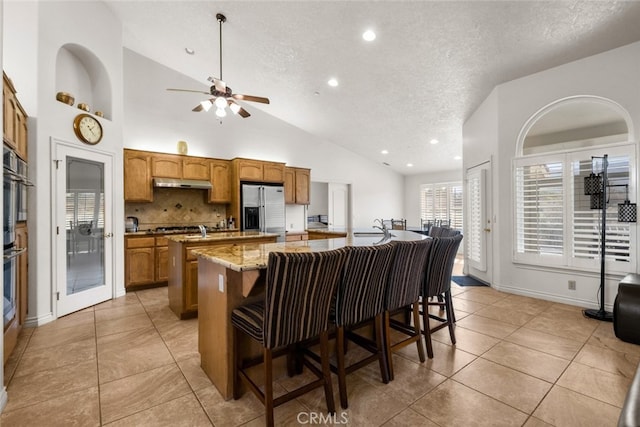 kitchen with a kitchen breakfast bar, light stone counters, a center island with sink, stainless steel fridge with ice dispenser, and decorative backsplash