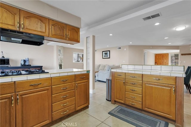 kitchen featuring light tile patterned floors, tile counters, kitchen peninsula, range hood, and black gas stovetop