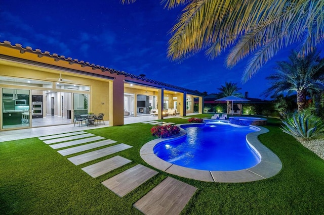 pool at night featuring an in ground hot tub, ceiling fan, a patio, and a lawn