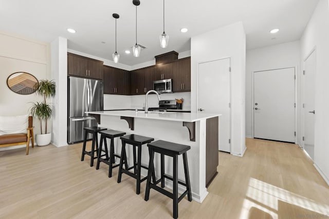 kitchen with pendant lighting, appliances with stainless steel finishes, a kitchen island with sink, dark brown cabinets, and light hardwood / wood-style floors