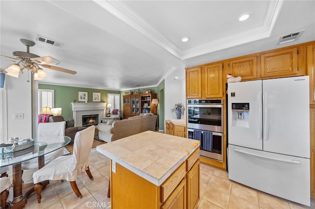 kitchen featuring a center island, white fridge with ice dispenser, a brick fireplace, a raised ceiling, and stainless steel double oven