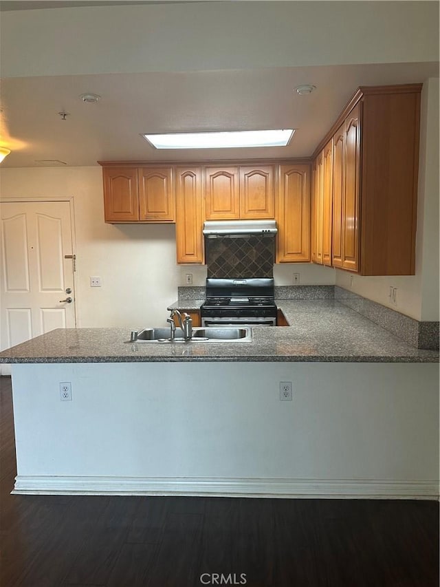 kitchen featuring dark stone counters, a peninsula, a sink, under cabinet range hood, and range with gas cooktop