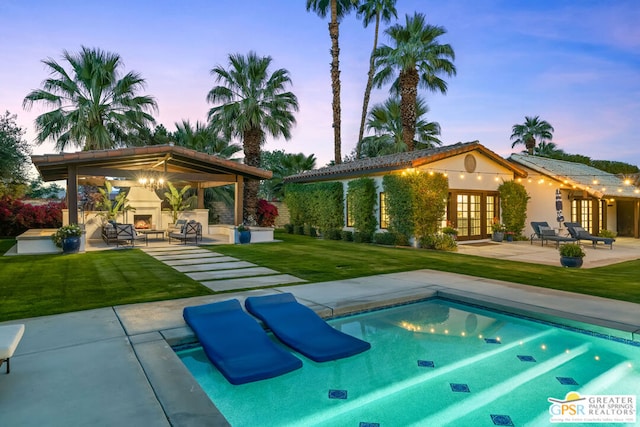 pool at dusk with a patio, exterior fireplace, a lawn, an outbuilding, and french doors