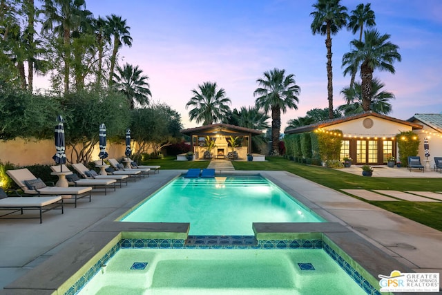 pool at dusk with an in ground hot tub, an outdoor structure, and a patio area