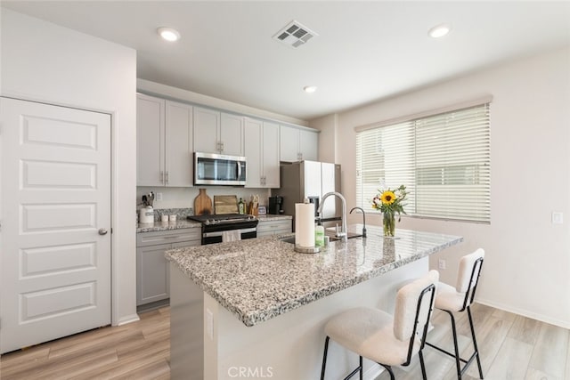 kitchen with light stone counters, stainless steel appliances, gray cabinets, and an island with sink