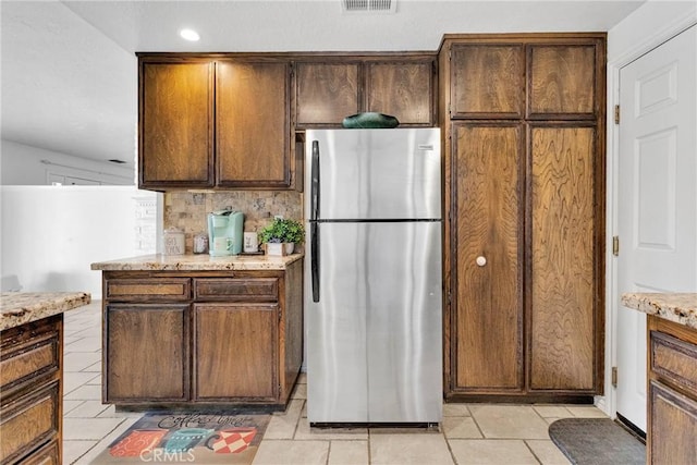 kitchen with stainless steel refrigerator, backsplash, light tile patterned floors, dark brown cabinetry, and light stone countertops