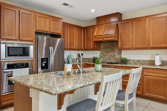 kitchen featuring sink, stainless steel appliances, light stone counters, an island with sink, and a kitchen bar