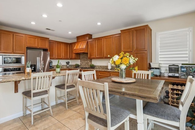 kitchen featuring light tile patterned floors, a breakfast bar area, appliances with stainless steel finishes, light stone countertops, and custom range hood