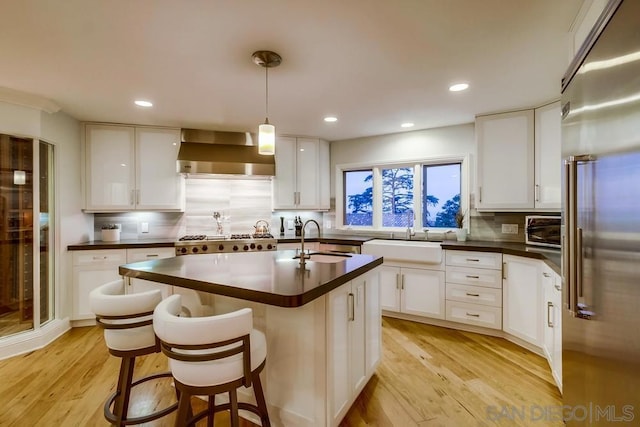 kitchen featuring white cabinets, stainless steel appliances, an island with sink, and wall chimney range hood