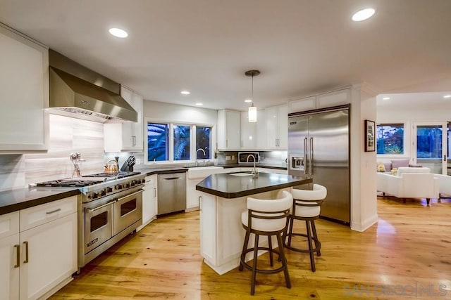 kitchen featuring wall chimney range hood, light hardwood / wood-style flooring, white cabinetry, high end appliances, and decorative light fixtures