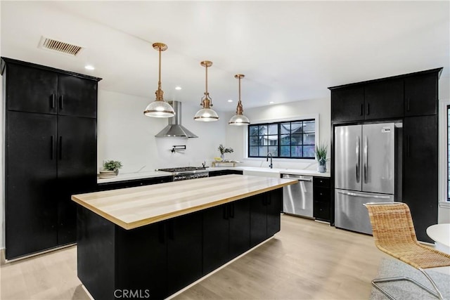 kitchen featuring wall chimney exhaust hood, appliances with stainless steel finishes, a kitchen island, pendant lighting, and light hardwood / wood-style floors