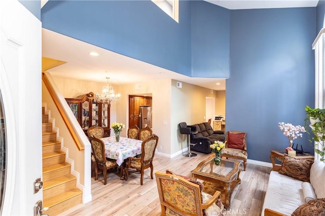 living room featuring a towering ceiling, stairway, an inviting chandelier, light wood-style floors, and baseboards