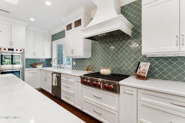 kitchen with sink, appliances with stainless steel finishes, light stone counters, custom range hood, and white cabinets