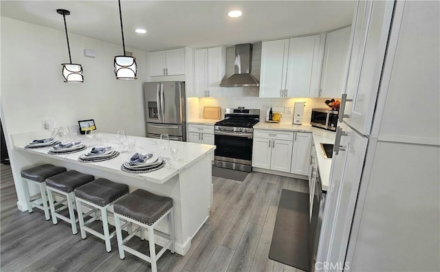 kitchen featuring wall chimney exhaust hood, white cabinetry, appliances with stainless steel finishes, a kitchen breakfast bar, and pendant lighting