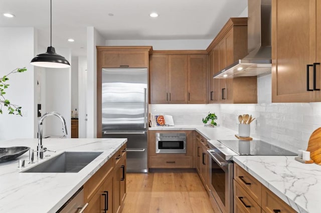 kitchen with sink, stainless steel appliances, light stone counters, decorative light fixtures, and wall chimney exhaust hood