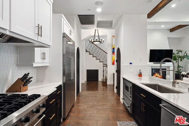 kitchen with sink, light stone counters, built in appliances, beamed ceiling, and white cabinets