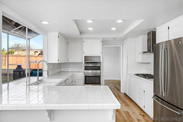 kitchen with sink, appliances with stainless steel finishes, tile counters, white cabinets, and wall chimney exhaust hood