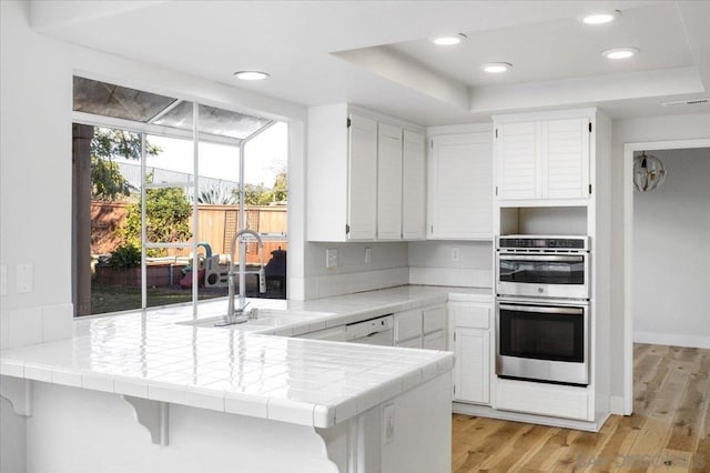 kitchen featuring tile countertops, double oven, sink, white cabinets, and kitchen peninsula