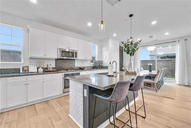 kitchen featuring dark stone countertops, hanging light fixtures, white cabinetry, stainless steel appliances, and a center island with sink