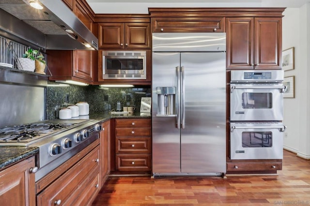 kitchen with range hood, dark hardwood / wood-style floors, built in appliances, tasteful backsplash, and dark stone counters