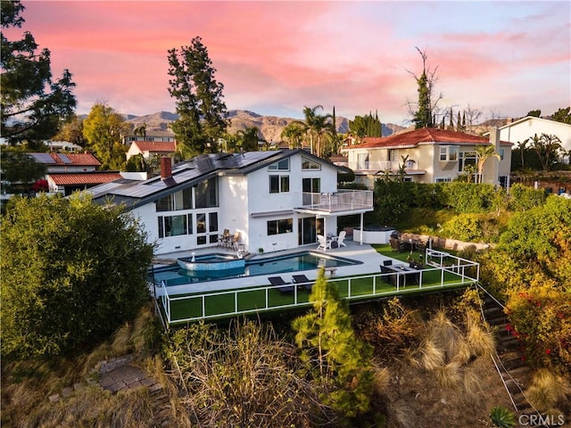 back house at dusk featuring a balcony, a swimming pool with hot tub, a mountain view, and a patio