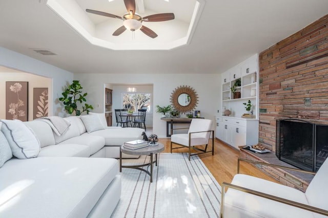 living room with a stone fireplace, built in features, ceiling fan, a tray ceiling, and light wood-type flooring