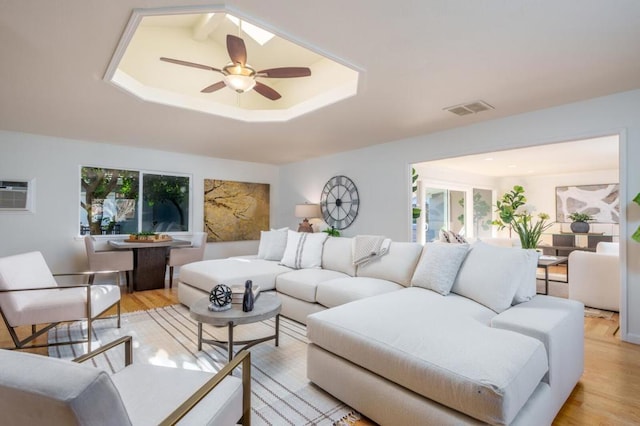 living room featuring a wall unit AC, light hardwood / wood-style floors, a raised ceiling, and ceiling fan